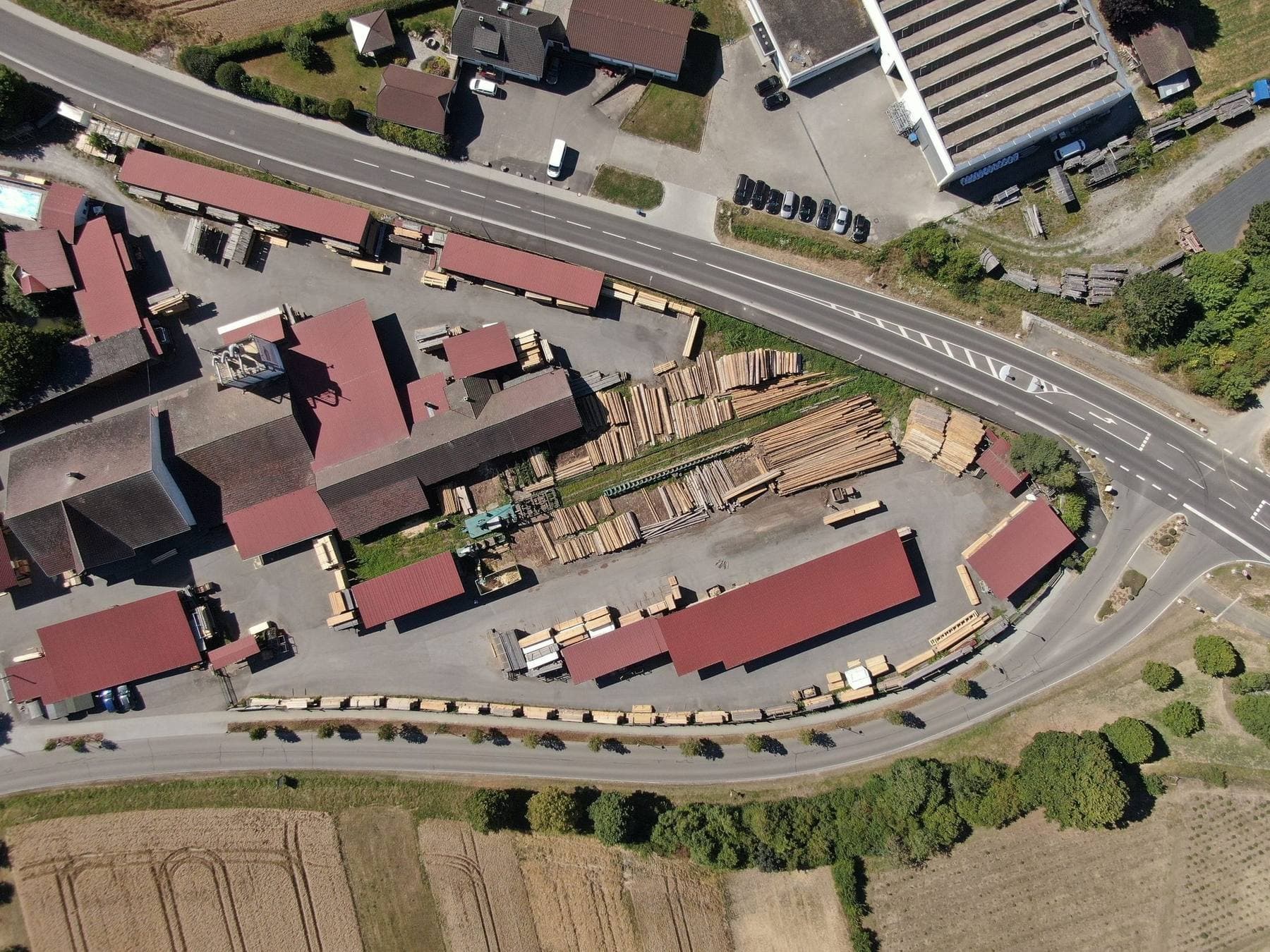 Luftaufnahme des Betriebsgeländes mit Hallen, Hof und Holzlager entlang der Straße
